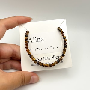 May include: A beaded bracelet featuring brown tiger eye gemstone beads and small gold-toned metal spacers. The bracelet is displayed on a white card printed with the name Alina, Morse code dots and dashes, and the text Muza Jewellery.