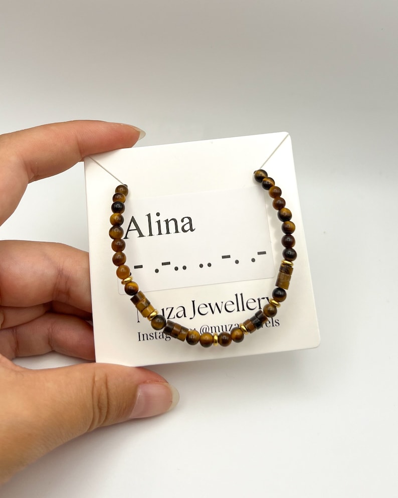 May include: A beaded bracelet featuring brown tiger eye gemstone beads and small gold-toned metal spacers. The bracelet is displayed on a white card printed with the name Alina, Morse code dots and dashes, and the text Muza Jewellery.