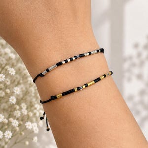 Può includere: Due braccialetti in cordino regolabili con piccole perline. Il braccialetto superiore presenta perline nere e color argento, mentre quello inferiore ha perline nere e color oro. Entrambi sono fissati con cordini neri sottili e nodi scorrevoli.