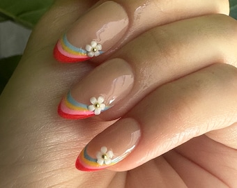 Puede incluir: Uñas en forma de almendra con base nude y punta francesa de colores arcoíris con un acento de flor blanca.