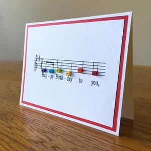 May include: A white card with a red border features a musical staff with the words "Happy Birthday to you" written on it. The words are spelled out using colorful beads.