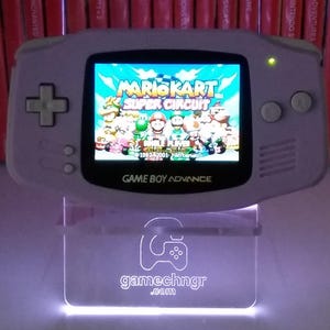 May include: A light grey Game Boy Advance console playing Mario Kart: Super Circuit. The console is on a clear, illuminated stand with "gamechngr.com". The background shows a shelf of red game cases.