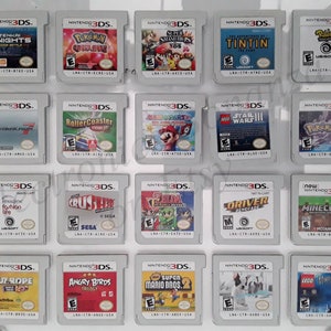 Puede incluir: Una colección de 18 cartuchos de juego de Nintendo 3DS que se muestran en un estante blanco. Los cartuchos están dispuestos en tres filas de seis. Los cartuchos son de diferentes colores y tienen diferentes títulos de juegos impresos en ellos. Algunos de los títulos incluyen "Super Smash Bros. 3DS", "Angry Birds Trilogy", "Minecraft" y "Lego Harry Potter".