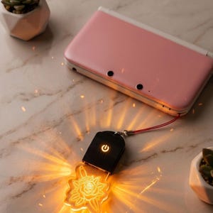 May include: A pink handheld games console with a white top sits beside a glowing black and clear acrylic charm. The charm features a rose and dagger design and an illuminated power button. The charm is connected to the console with a red cord.