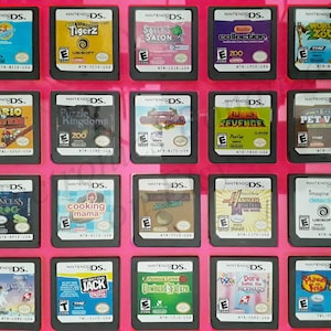 Puede incluir: Una colección de 18 cartuchos de juego de Nintendo DS que se muestran en una caja rosa. Los cartuchos están dispuestos en una cuadrícula de tres filas y seis columnas. Los cartuchos son de diferentes colores y tienen diferentes títulos, que incluyen "Tigerz", "Sally's Salon", "Puzzle Kingdoms", "Mario Kart DS", "Cooking Mama", "Princess Frog", "Hannah Montana", "Pet Vet", "Dora Saves the Mermaid", "Jack  Tonic", "Unwound Future", "World Zoo", "Imagine Detective" y "The Princess and the Frog".