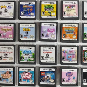 May include: A collection of 18 Nintendo DS game cartridges, displayed in a clear plastic case. The cartridges are arranged in three rows of six, with each cartridge featuring a different game title and cover art. Some of the games include "Tangled", "Catz", "Plants vs. Zombies", "Dora the Explorer", "Cooking Mama", and "Big Brain Academy".