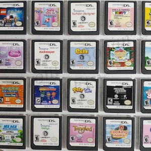 May include: A collection of 18 Nintendo DS game cartridges, displayed in a clear plastic case. The cartridges are arranged in three rows of six, with each cartridge featuring a different game title and cover art. Some of the games include "Dora Saves the Mermaids", "Imagine Fashion Designer", "Hello Kitty Party", "Plants vs. Zombies", "Disney Princess", "Imagine Zookeeper", "Catz", "Gardening Mama", "Dora Puppy", "Team Umizoomi", "Petz Catz Clan", "Michael Jackson Experience", "Big Brain Academy", "Her Ace", "Imagine Babyz", "Tangled", "Cooking Mama", and "My Little Pony: Friendship is Magic".