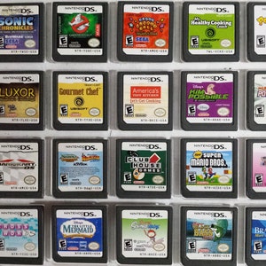 Op de afbeelding: Een verzameling van 18 Nintendo DS-spelcartridges die worden getoond in een doorzichtige plastic doos. De cartridges zijn gerangschikt in drie rijen van zes. De cartridges zijn allemaal in verschillende kleuren en hebben verschillende titels, waaronder "Sonic Chronicles", "Gourmet Chef", "Mario Kart DS" en "Brain Age 2".
