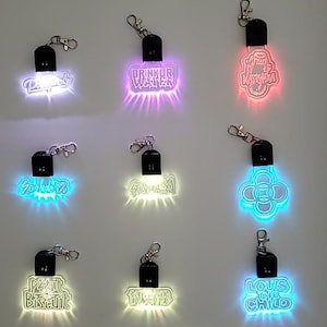 May include: A collection of illuminated keychains with various designs and text, including "Drinkur Water" and "Louis the Child." Each keychain has a black top and a silver clasp. The keychains emit different colors, such as blue, pink, and yellow.
