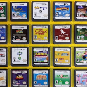 May include: A collection of 18 Nintendo DS game cartridges displayed in a yellow plastic case. The cartridges are arranged in a grid of 3 rows and 6 columns. The cartridges are all different colors and feature various characters and logos. Some of the games include: Club House Games, Tinker Bell, Petz Nursery 2, Madagascar 3, Food Frenzy, Cosmic Destruction, Quest for Go, The Simpsons Game, Nintendogs, Tinker Bell, Mario Kart DS, Sims 3, Sonic Classic Collection, Treasure Adventure, Go Pets, Hannah Montana Music Jam, CN Racing, Naruto Ninja Destiny, Jigapix, and Dora Saves the Mermaids.