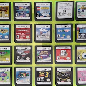 Könnte beinhalten: Eine Sammlung von 18 Nintendo DS-Spielkassetten, die in einem durchsichtigen Plastikbehälter auf einem leuchtend grünen Hintergrund ausgestellt sind. Die Kassetten sind in einem Raster von 3 Reihen und 6 Spalten angeordnet. Die Kassetten sind alle in verschiedenen Farben und zeigen eine Vielzahl beliebter Videospieltitel, darunter "Hannah Montana", "iCarly 2", "Lego Batman", "Up", "Toy Story 3" und "Math Blaster".