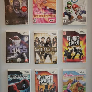 Może przedstawiać: Dziewięć pudełek z grami na Nintendo Wii z różnymi tytułami, w tym "Gdzie mieszkają dzikie zwierzęta", "Barbie Jet Set & Style", "Mario Kart Wii", "The Bigs", "Sing It Party Hits", "Guitar Hero World Tour", "Spider-Man Edge of Time", "Guitar Hero III Legends of Rock" i "Barbie Horse Adventures Riding Camp".