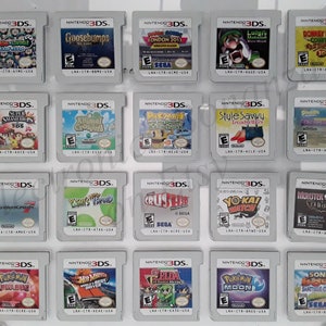 Puede incluir: Una colección de cartuchos de juegos de Nintendo 3DS, con títulos como "Mario Kart 7", "Goosebumps" y "Pokemon Moon". Los cartuchos se muestran sobre una superficie transparente, mostrando los títulos y las ilustraciones.