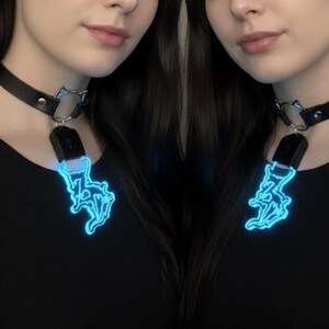 May include: Black leather choker necklace with a silver-tone cat ear charm and a glowing blue pendant. The pendant is a stylized lightning bolt design. The necklace is worn against a black top.