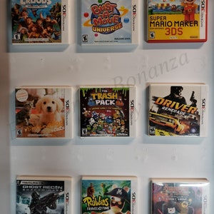 Könnte beinhalten: Neun Nintendo 3DS-Spielkartuschen, die an einer weißen Wand ausgestellt sind. Die Spiele sind The Croods: Prehistoric Party, Bust-A-Move Universe, Super Mario Maker 3DS, Golden Retriever + Coats, The Trash Pack, Driver Renegade, Tom Clancy's Ghost Recon: Shadow Wars, Rabbids Travel in Time und Adventure Time: I Don't Know.