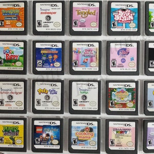May include: A collection of 18 Nintendo DS game cartridges, displayed in a clear plastic case. The cartridges are arranged in three rows of six, with each cartridge featuring a different game title and cover art. Some of the games include "Imagine Zookeeper", "Tangled", "Dora the Explorer", "Plants vs. Zombies", and "Cooking Mama".