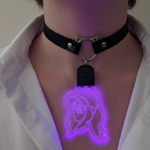 Pode incluir: Colar gargantilha de couro preto com um pingente de orelha de gato prateado. Um pingente de néon roxo em forma de mulher com óculos e chapéu pende do colar. O pingente é iluminado por uma luz roxa brilhante.