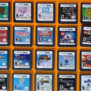 Puede incluir: Una colección de 20 cartuchos de juegos de Nintendo DS que se muestran en una caja de plástico transparente con un fondo naranja brillante. Los cartuchos están organizados en cuatro filas de cinco, con cada cartucho presentando un título de juego y una portada diferentes. Algunos de los juegos incluyen Animal Crossing: Wild World, Hello Kitty: Big City Dreams, MySims Kingdom, LEGO Harry Potter: Years 1-4 y LEGO Star Wars: The Complete Saga.