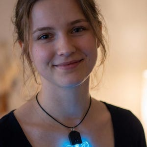 Op de afbeelding: Een zwarte ketting met een blauwe, oplichtende hanger in de vorm van een knipogende cartoon wasbeer. De hanger wordt verlicht door een helderblauw licht. De persoon die de ketting draagt, glimlacht.