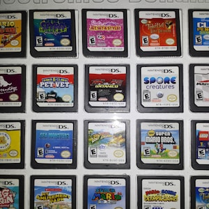 May include: A collection of 18 Nintendo DS game cartridges, including popular titles like Mario Kart DS, Super Mario Bros., and  Tinker Bell and the Lost Treasure. The cartridges are arranged in a grid pattern on a white surface.