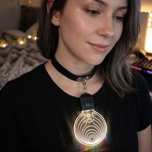 Puede incluir: Collar gargantilla negro con un colgante luminoso con forma de bombilla. El colgante circular presenta líneas concéntricas y emite una luz de arcoíris. El collar se lleva con una camiseta negra.