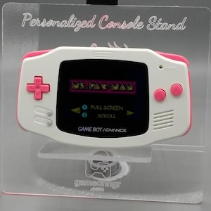 Peut inclure: Console de jeu Game Boy Advance blanche et rose affichant le jeu Ms. Pac-Man. La console est présentée sur un support transparent avec le texte "Personalized Console Stand" au-dessus. L'écran affiche les options "Full Screen" et "Scroll".