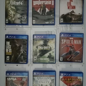 May include: A wall display of nine PlayStation 4 video game cases. The cases are arranged in three rows of three. The games include Fallout 4, Wolfenstein II, The Evil Within, The Last of Us Remastered, Call of Duty: Infinite Warfare, Spider-Man, NBA 2K15, Assassin's Creed Unity, and God of War Remastered.