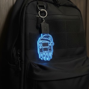 May include: A glowing blue keychain featuring a video game console design with a Santa hat and icicles. The keychain is attached to a black backpack with a silver key ring.