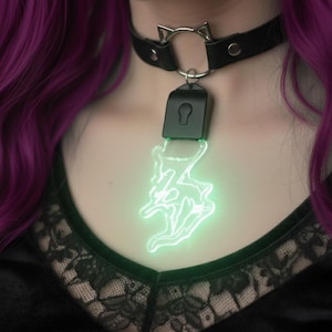 Puede incluir: Collar gargantilla de cuero negro con un anillo plateado con forma de orejas de gato y un colgante verde brillante. El colgante presenta un diseño de ojo de cerradura y una figura de murciélago verde neón. El collar se asegura con una hebilla.