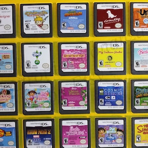 Puede incluir: Una colección de 18 cartuchos de juego de Nintendo DS, expuestos en una caja de plástico amarilla. Los cartuchos están dispuestos en tres filas de seis, con cada cartucho presentando un título de juego y una ilustración de portada diferentes. Algunos de los juegos incluyen "Imagine Fashion Designer", "Barbie Jet Set & Style", "Cooking Mama", "Iron Man 2" y "The Simpsons Game".