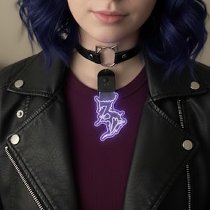 May include: Black leather choker necklace with a silver cat ear charm and a purple neon cat pendant. The pendant is illuminated and hangs from a black rectangular base. The person is wearing a black leather jacket and a burgundy shirt.
