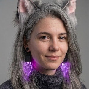 Può includere: Una persona indossa una fascia per capelli con orecchie di lupo in finta pelliccia grigia e orecchini neri con un disegno di impronta di zampa che si illumina di luce viola. Indossa un maglione grigio scuro e ha lunghi capelli grigi.