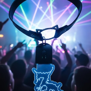 Puede incluir: Collar de cuero negro con diseño de orejas de gato y un colgante azul brillante. El colgante presenta una silueta de gato estilizada. El fondo muestra una multitud de personas y luces láser coloridas.