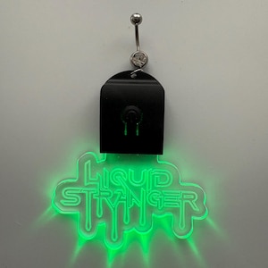 Puede incluir: Un piercing para el ombligo con una parte superior negra y un logotipo "Liquid Stranger" verde brillante. El logotipo está en una fuente que gotea, y el anillo tiene una barra plateada y una gema transparente.