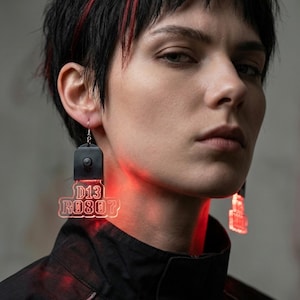 May include: Black earrings with the text "D13 ROB07" in red neon. The earrings are rectangular with a small black top piece. The person in the image is wearing a black jacket and has black hair with red highlights.