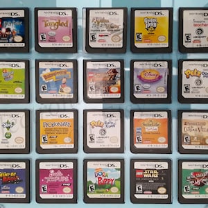Peut inclure: Une collection de 16 cartouches de jeu Nintendo DS présentées dans un boîtier en plastique transparent. Les cartouches sont disposées en quatre rangées de quatre, chaque cartouche présentant un titre de jeu et une illustration de couverture différents. Parmi les jeux, on trouve "Tangled", "Guitar Hero", "Lego Harry Potter", "Sims", "Dora", "Lego Star Wars" et "Suite Life".