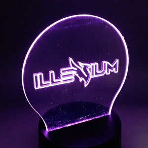 Peut inclure: Une veilleuse LED violette avec une base en acrylique transparente représentant le mot "ILLENIUM" en lettres blanches avec un graphisme d'oiseau stylisé.