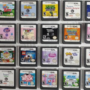 May include: A collection of 18 Nintendo DS game cartridges, displayed in a clear plastic case. The cartridges are arranged in three rows of six, with each cartridge featuring a different game title and cover art. Some of the games include Dora the Explorer, Hello Kitty, Plants vs. Zombies, and Tangled.