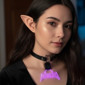Pode incluir: Uma pessoa usando orelhas de elfo e um colar gargantilha preto com fecho em forma de gato. O colar apresenta um pingente roxo brilhante com a palavra "MARAUDA" em uma fonte estilizada.