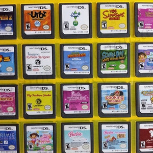 Puede incluir: Una colección de 18 cartuchos de juego de Nintendo DS, expuestos sobre un fondo amarillo. Los cartuchos presentan una variedad de juegos populares, incluyendo Iron Man 2, The Urbz, Sims 3, The Simpsons Game, Island Princess, Phineas y Ferb, Imagine Fashion Designer, Super Slam, Scooby-Doo! ¿Quién está mirando a quién?, Refugio de mascotas, Jet Set & Style, My Fashion Studio, Barbie Puedo ser... Cachorros, Cooking Mama, El amanecer de la vida, Destrucción cósmica, Dora la Exploradora, Barbie, Barbie y Nintendogs.