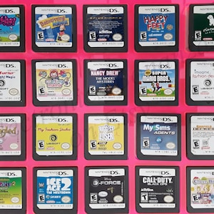 Puede incluir: Una colección de cartuchos de juegos de Nintendo DS, dispuestos sobre una superficie rosa. Los títulos incluyen "Zombies!", "Babysitting Mania", "Happy Feet", "Super Mario Bros." y "Call of Duty: Black Ops". Los juegos están en carcasas negras.