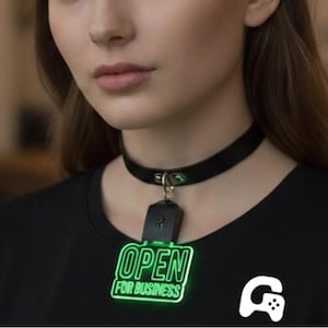 Peut inclure: Un collier ras du cou noir avec un fermoir argenté et un pendentif vert néon indiquant "OPEN FOR BUSINESS". Le pendentif est attaché à un porte-clés noir. Un t-shirt noir avec un logo blanc est également visible.
