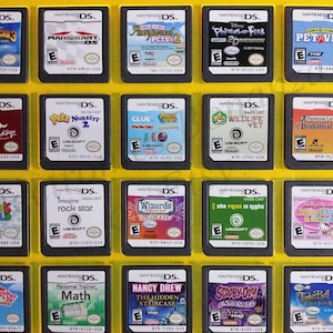 May include: A collection of 18 Nintendo DS game cartridges, displayed in a yellow frame. The cartridges are arranged in three rows of six, with each cartridge featuring a different game title and cover art. Some of the games include Madagascar 3, Mario Kart DS,  and Hello Kitty.