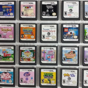 May include: A collection of 18 Nintendo DS game cartridges, displayed in a grid. The cartridges are in various colors and feature different game titles, including "Tangled", "Dora", "Plants vs. Zombies", and "Cooking Mama".
