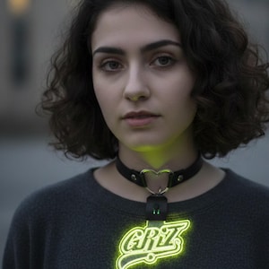 Pode incluir: Um colar gargantilha de couro preto com um pingente em forma de coração e um logotipo GRiZ verde neon brilhante. O colar é usado sobre um suéter cinza escuro.