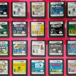 Puede incluir: Una colección de 16 cartuchos de juegos de Nintendo DS que se muestran en una caja de plástico rosa. Los cartuchos están dispuestos en cuatro filas de cuatro. Los juegos incluyen títulos como "Super Mario Bros.", "Nancy Drew", "My Sims Agents", "iCarly 2", "Happy Feet", "Ice Age 2" y "Enredados".