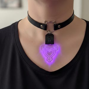 Pode incluir: Colar gargantilha de couro preto com um pingente de orelha de gato prateado e um pingente de símbolo transgênero roxo brilhante. O pingente é um coração estilizado com símbolos masculinos e femininos.