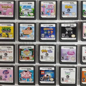 May include: A collection of 18 Nintendo DS game cartridges, displayed in a black plastic case. The cartridges are arranged in three rows of six, with each cartridge featuring a different game title and cover art. The games include titles such as "Tangled", "Plants vs. Zombies", "Cooking Mama", "Dora the Explorer", "Catz", "Hello Kitty", "Imagine Fashion Designer", "Gardening Mama", "Petz Catz Clan", "Disney Princess", "Zookeeper", "Big Brain Academy", "Lego Harry Potter", "Imagine Babyz", and "Tinkerbell and the Lost Treasure".