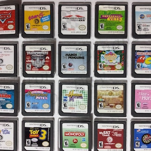 Op de afbeelding: Een verzameling van 15 Nintendo DS-spelcartridges, waaronder titels als "Cars", "Transformers Autobots", "De Mars van de Pinguïns", "Hannah Montana" en "Toy Story 3".