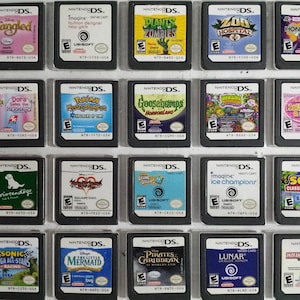 May include: A collection of Nintendo DS game cartridges, featuring titles like "Tangled," "Plants vs. Zombies," and "Sonic All-Stars Racing." The games have colorful labels with various game art and logos. The cartridges are displayed on a white surface.